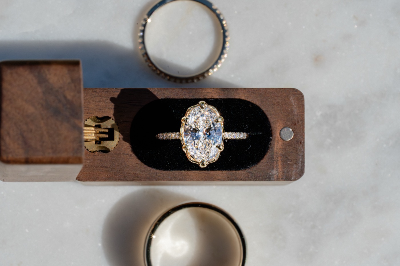 An oval diamond solitaire in a walnut ring box, with two bands beside it.